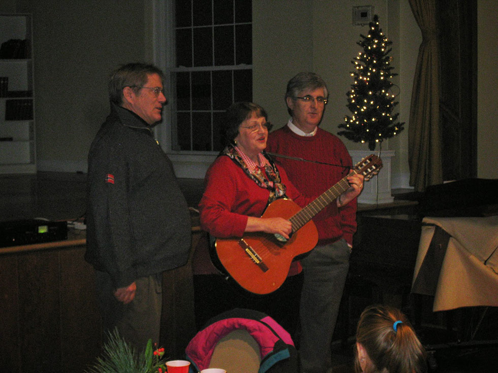 Cheri Christakis playing a guitar and singing with two men in the church